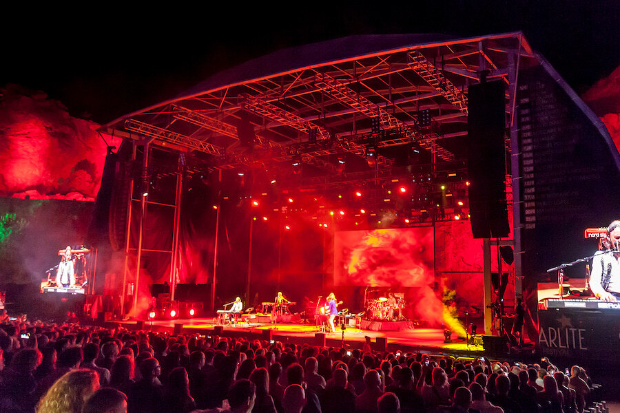 Marbellas besten Sommerfestivals. Open-Air-Konzert bei Nacht mit einer großen Menschenmenge vor einer hell erleuchteten Bühne mit Live-Musikern und leuchtend roten Lichteffekten vor einer dramatischen Rockkulisse.
