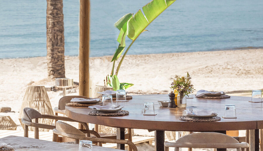 Outdoor beachside dining area at Puente Romano chiringuito, featuring a large round wooden table set with plates, glasses, and cutlery, surrounded by wooden chairs, with a sandy beach and calm sea in the background.