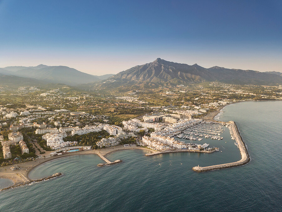 Vista aérea de Puerto Banús y los alrededores de Marbella, España, con el mar Mediterráneo en primer plano y la montaña La Concha dominando el fondo bajo un cielo azul claro.