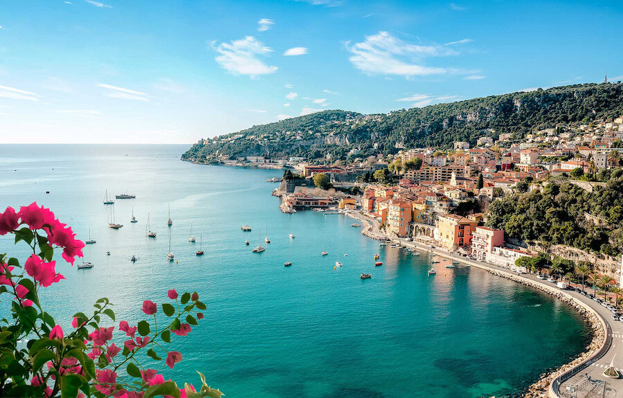 Investeerders verkiezen Marbella boven de Franse Rivièra. Een schilderachtig uitzicht op de Franse Rivièra met turquoise wateren, kleurrijke gebouwen aan zee, jachten die voor anker liggen in de baai en felroze bougainvillea op de voorgrond. Dit alles wordt omlijst door groene heuvels onder een helderblauwe lucht.