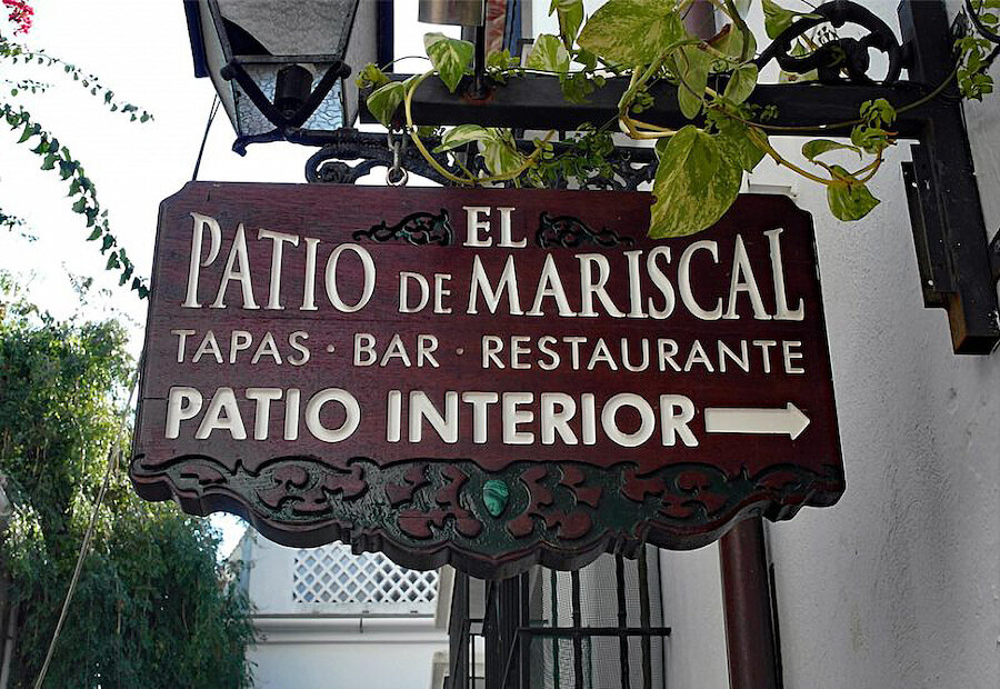 Wooden hanging sign for El Patio de Mariscal restaurant, a tapas bar and restaurant, indicating the direction to the interior patio, mounted on a white wall with greenery around.