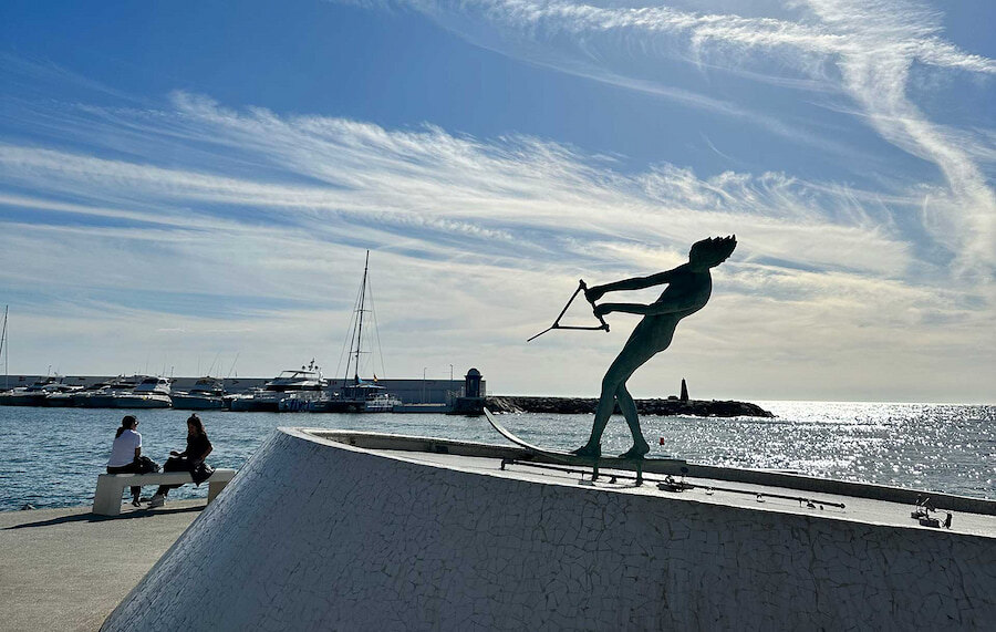 Bronzen beeld van een waterskiër met een sleepkabel, dramatisch zwevend boven een gebogen mozaïekvoetstuk aan de kust van Marbella. Op de achtergrond zijn jachten, een vuurtoren en twee personen op een bankje te zien, met uitzicht op de glinsterende Middellandse Zee.
