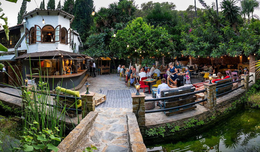 Outdoor dining area at Le Patio by Cascada Marbella, featuring guests seated at colorful tables surrounded by lush greenery, string lights, and a stone bridge over a small pond in the foreground.