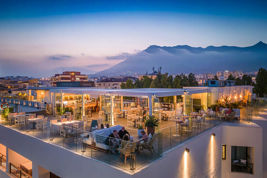 Evening view of Edge by Paco Pérez restaurant rooftop terrace, with modern outdoor seating, diners enjoying their meals, glass railings, and a backdrop of mountains and city lights under a twilight sky