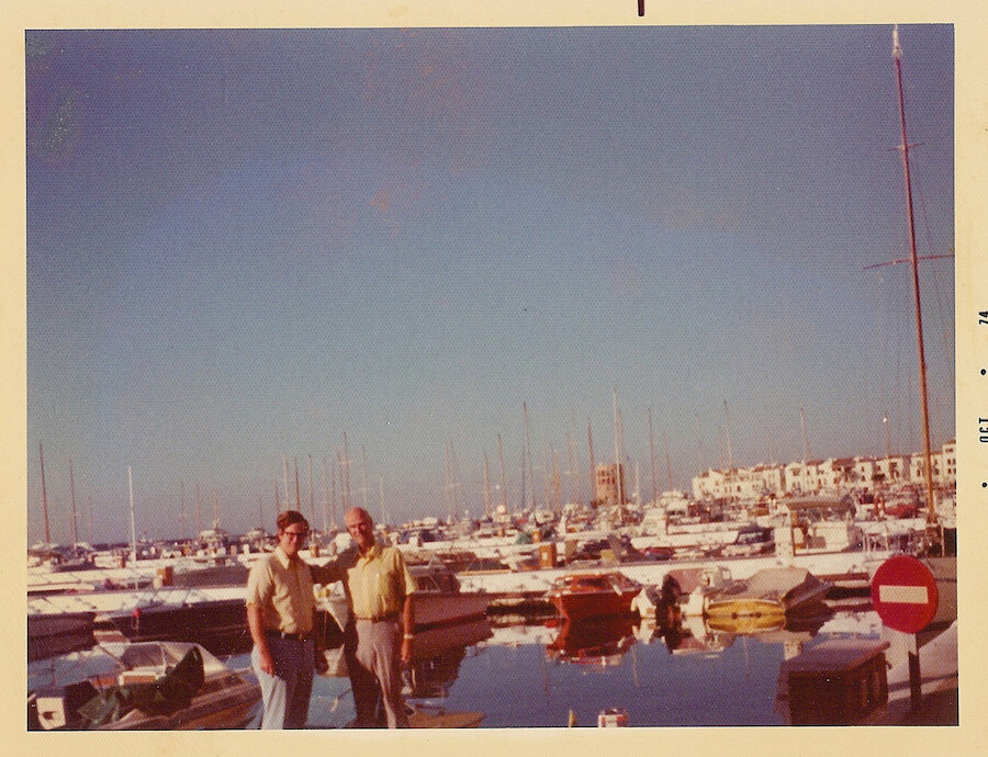 Photo d'époque de deux hommes debout dans une marina remplie de voiliers et de yachts, avec des bâtiments blancs de style méditerranéen en arrière-plan. Prise à Puerto Banús, Marbella, en octobre 1974.
