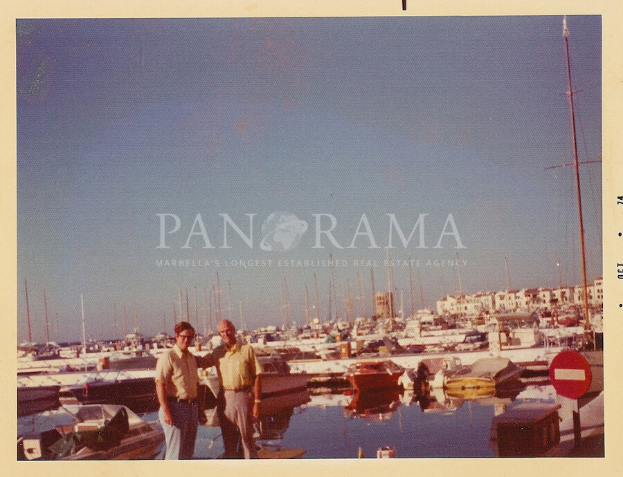 Vintage photo of two men standing at a marina filled with sailboats and yachts, with white Mediterranean-style buildings in the background. Taken in Puerto Banús, Marbella, dated October 1974