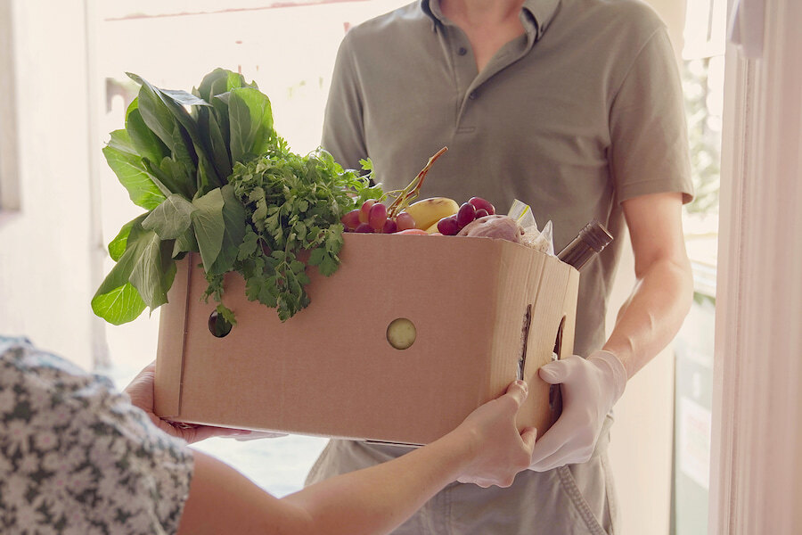 Een persoon ontvangt bij de deur een doos met verse boodschappen, waaronder bladgroenten, kruiden, fruit en een stokbrood, van een bezorger die handschoenen draagt.
