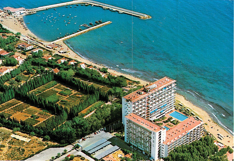 Vue aérienne d'un complexe hôtelier en bord de mer avec piscine, situé à côté d'une petite marina et d'un littoral sablonneux, entouré de jardins et de parcelles agricoles
