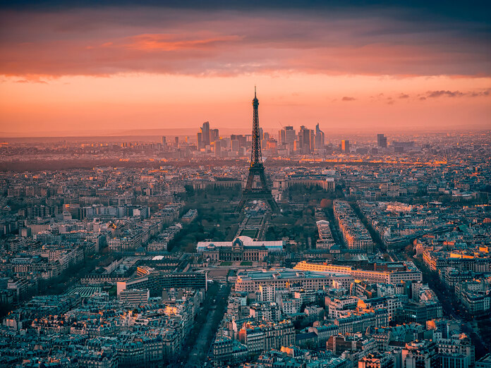 Luchtfoto van Parijs bij zonsondergang met de Eiffeltoren in het midden en de skyline van La Défense op de achtergrond, onder een dramatische hemel.
