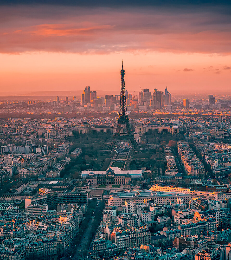 Vue aérienne de Paris au coucher du soleil avec la Tour Eiffel au centre et les toits de La Défense en arrière-plan sous un ciel dramatique.