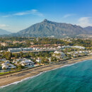 Aerial view of Marbella's Golden Mile, showcasing luxury beachfront villas, the Puente Romano resort, and the backdrop of La Concha mountain under a clear blue sky.