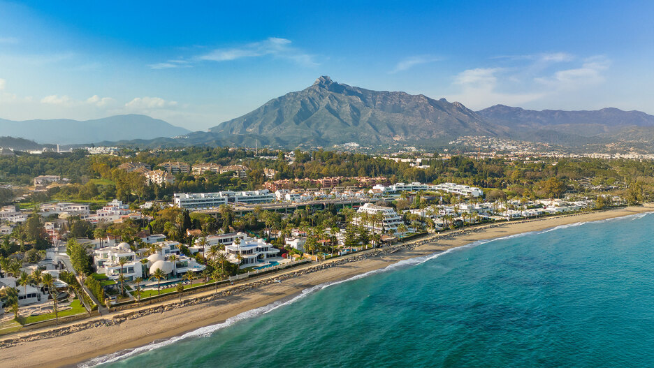 Luftaufnahme der Goldenen Meile von Marbella mit luxuriösen Strandvillen, dem Resort Puente Romano und dem Berg La Concha im Hintergrund unter einem klaren blauen Himmel.