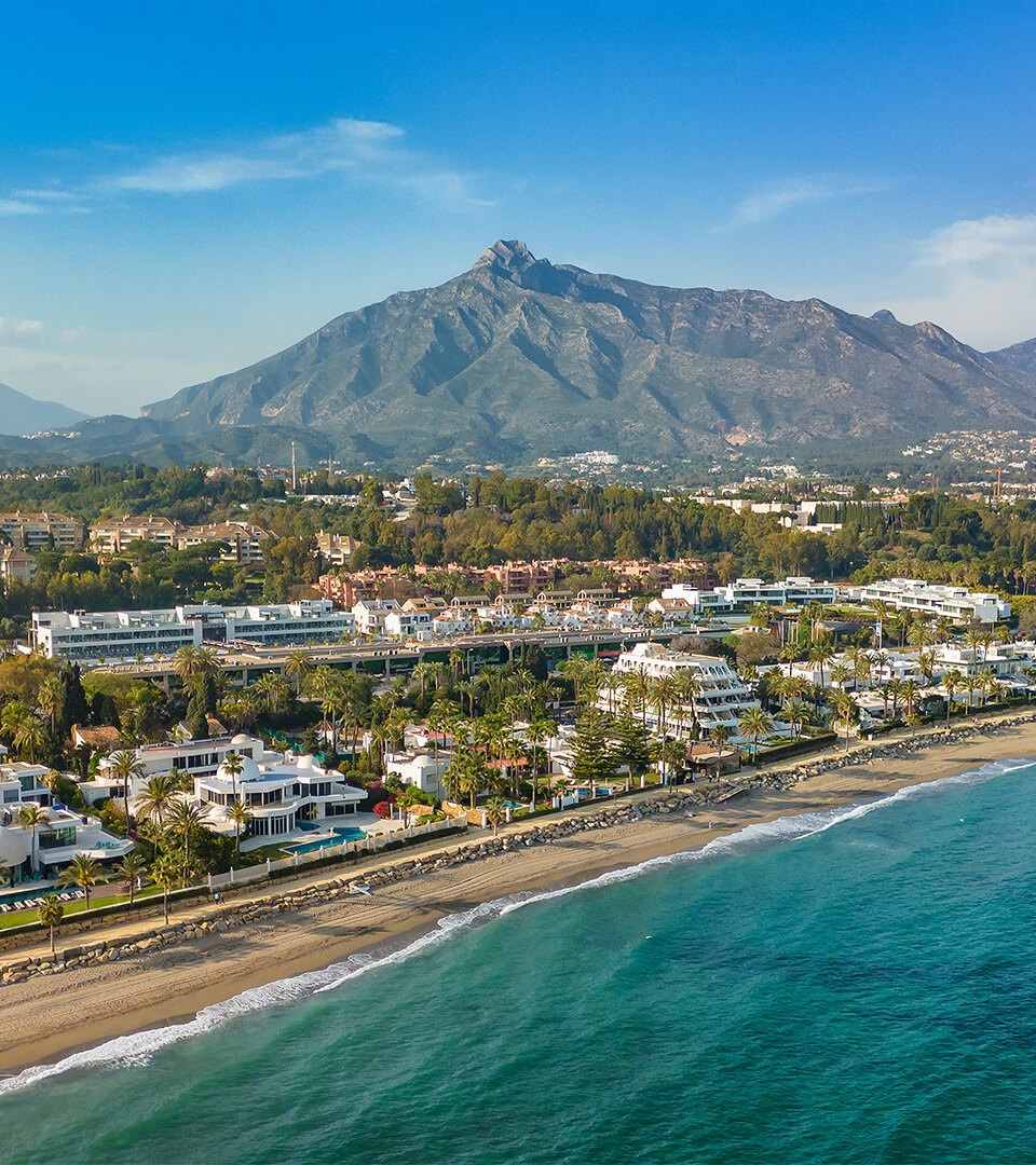 Vista aérea de la Milla de Oro de Marbella, que muestra lujosas villas frente al mar, el complejo Puente Romano y el telón de fondo de la montaña La Concha bajo un cielo azul claro.