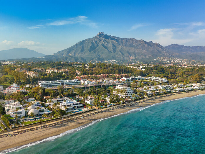Luchtfoto van de Golden Mile van Marbella, met luxe villa's aan het strand, het Puente Romano-resort en de berg La Concha op de achtergrond, onder een helderblauwe hemel.