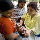 A healthcare worker administers oral vaccine drops to a baby held by a smiling mother during a public health campaign, with other people observing in the background.