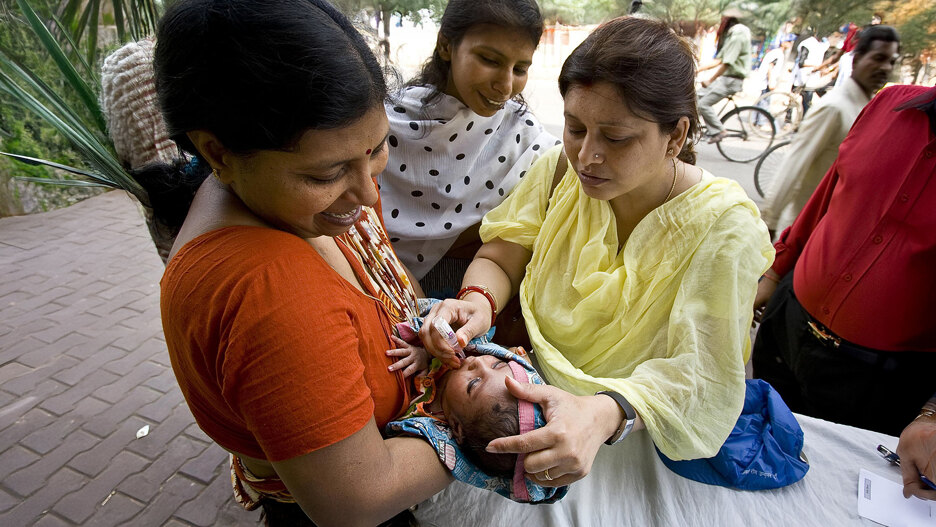 A healthcare worker administers oral vaccine drops to a baby held by a smiling mother during a public health campaign, with other people observing in the background.