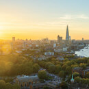 Adembenemend panoramisch uitzicht over Londen bij zonsondergang, met The Shard, de rivier de Theems, de London Eye en een stadsgezicht dat baadt in gouden licht, met een voorgrond van weelderige groene bomen en historische gebouwen.