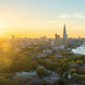 Stunning panoramic view of London at sunset featuring The Shard, the River Thames, the London Eye, and a cityscape bathed in golden light, with a foreground of lush green trees and historic buildings.