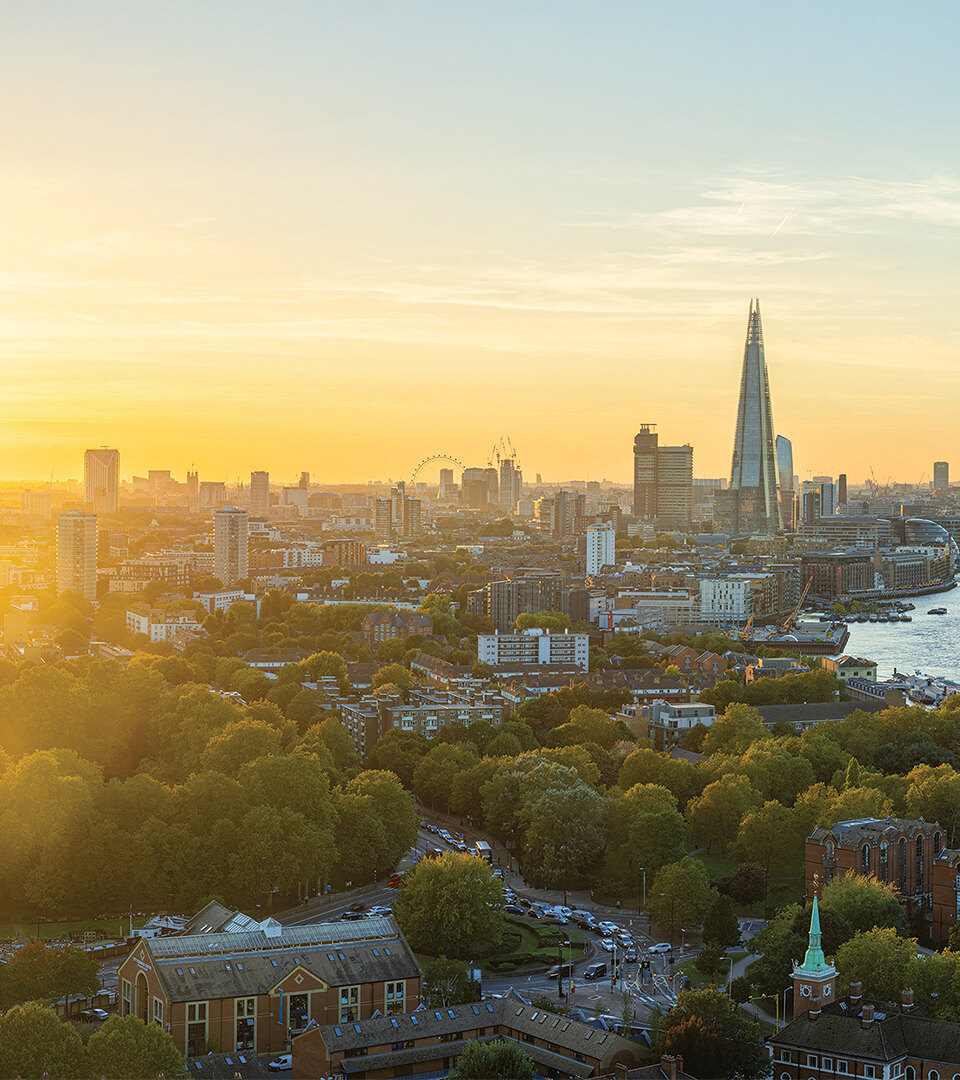 Stunning panoramic view of London at sunset featuring The Shard, the River Thames, the London Eye, and a cityscape bathed in golden light, with a foreground of lush green trees and historic buildings.