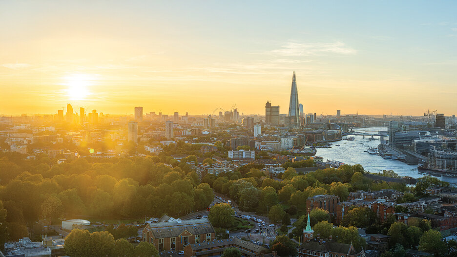 Impresionante vista panorámica de Londres al atardecer con The Shard, el río Támesis, el London Eye y un paisaje urbano bañado por una luz dorada, con un primer plano de exuberantes árboles verdes y edificios históricos.