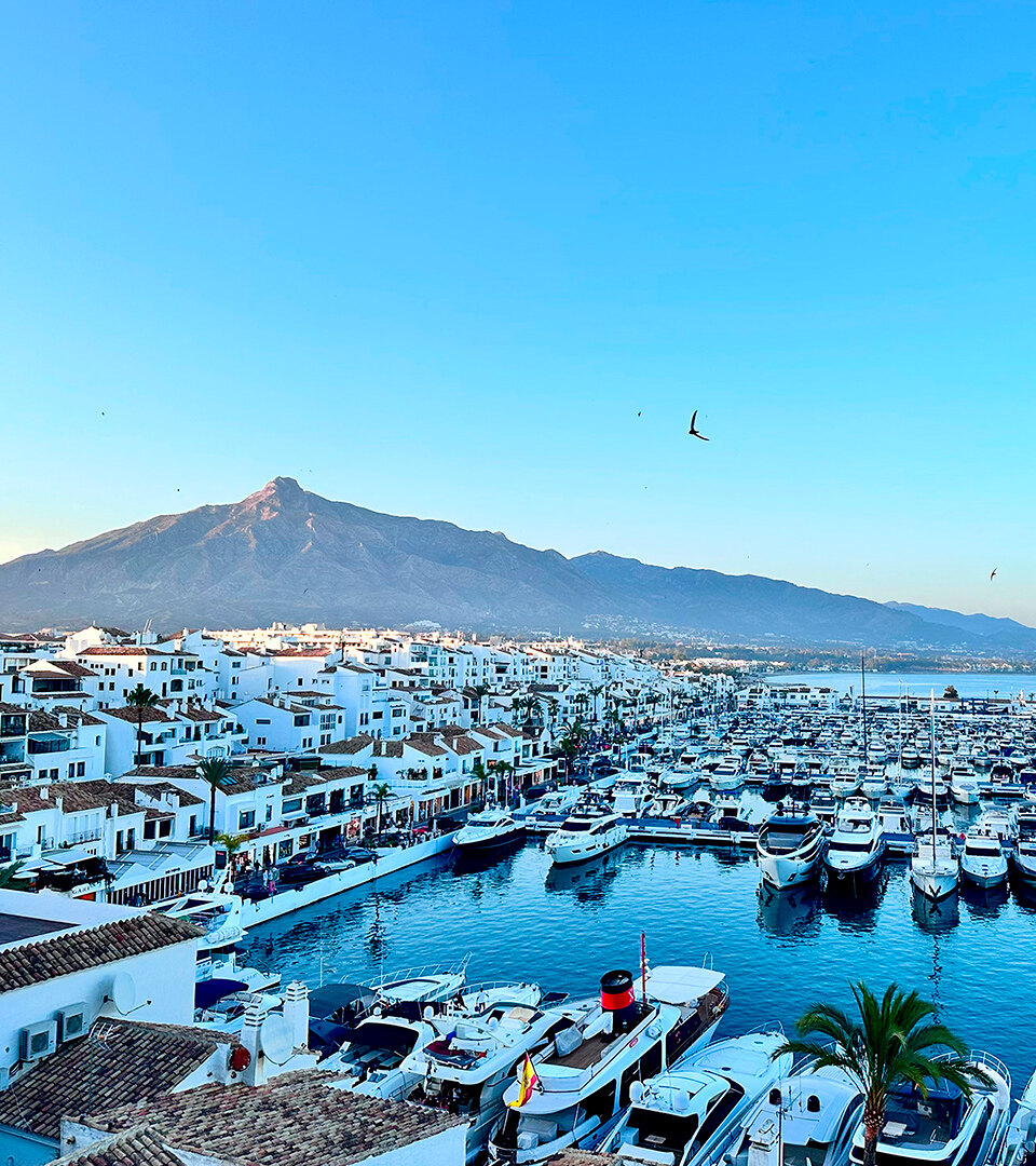 Panoramic view of Puerto Banús marina in Marbella, with rows of luxury yachts, whitewashed buildings, and the iconic La Concha mountain in the background. The clear blue sky and calm Mediterranean waters highlight the beauty of this exclusive coastal destination.