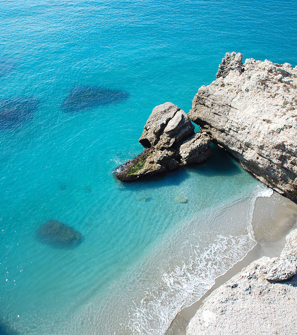 View from above of a small secluded beach with turquoise waters and striking rock formations. Waves gently lap against the shore, and submerged rocks are visible beneath the crystal-clear sea.
