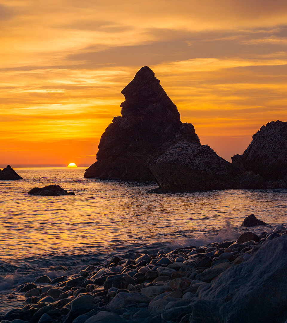 Dramatischer Sonnenuntergang über einem felsigen Strand in Málaga, mit zerklüfteten Felsnadeln, die sich vor einem leuchtend orangefarbenen und violetten Himmel abzeichnen, und Wellen, die sanft über glatte Steine ​​schwappen.
