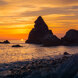 Dramatic sunset over a rocky beach in Málaga, with jagged sea stacks silhouetted against a glowing orange and purple sky, and waves gently washing over smooth stones.