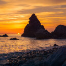 Dramatic sunset over a rocky beach in Málaga, with jagged sea stacks silhouetted against a glowing orange and purple sky, and waves gently washing over smooth stones.