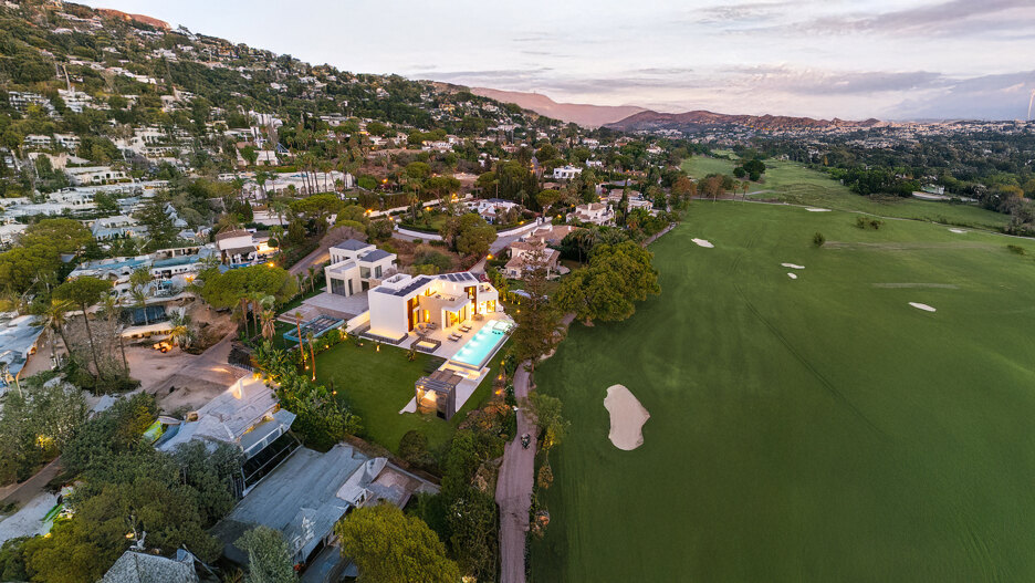 Aerial sunset view of a contemporary frontline golf villa in Marbella, illuminated against the lush green fairways and nestled at the foot of the hills, with panoramic mountain views and a tranquil residential setting.