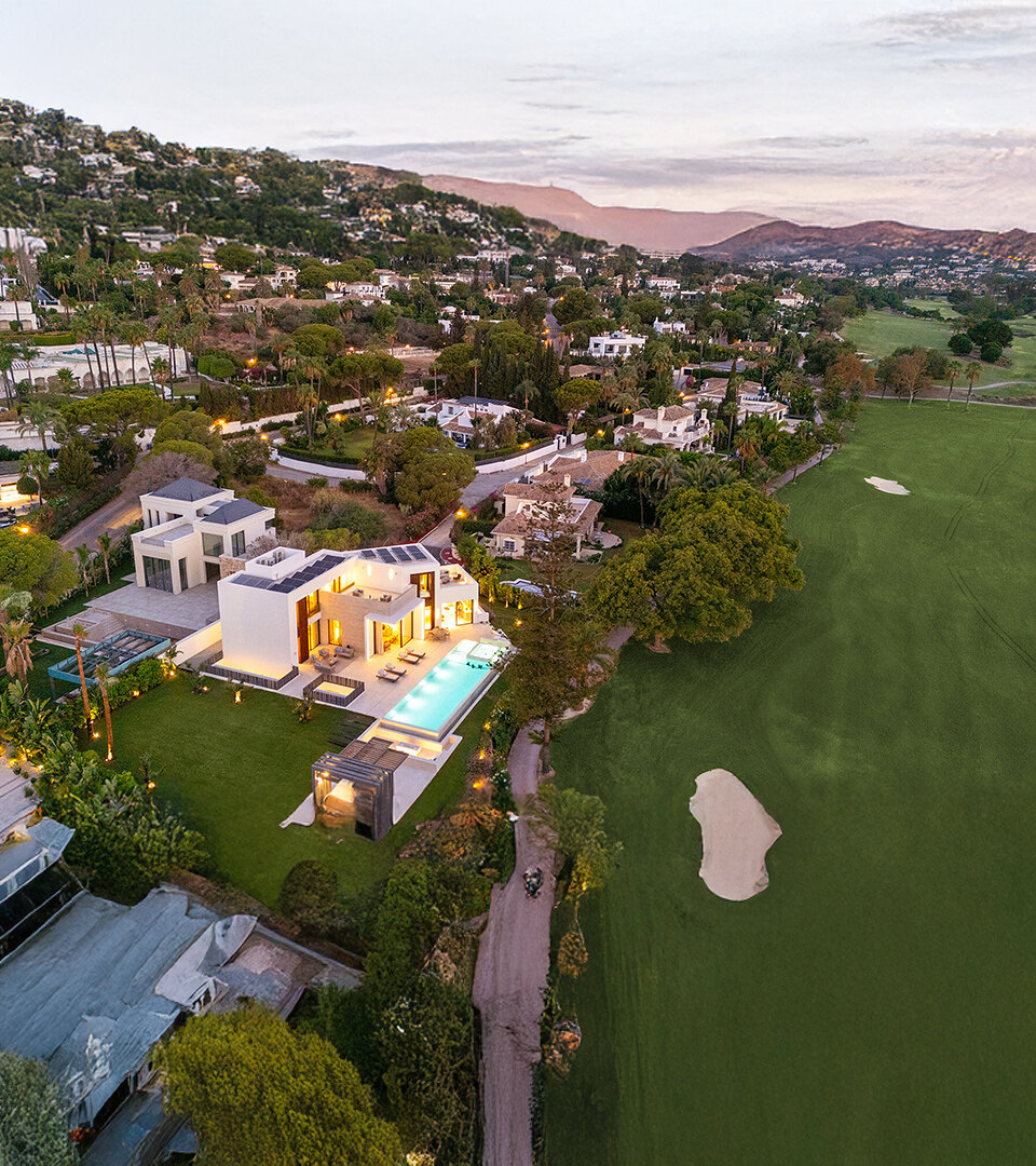 Vista aérea del atardecer de una villa contemporánea en primera línea de golf en Marbella, iluminada contra los exuberantes fairways verdes y ubicada al pie de las colinas, con vistas panorámicas a la montaña y un entorno residencial tranquilo.
