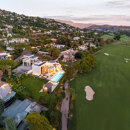 Aerial sunset view of a contemporary frontline golf villa in Marbella, illuminated against the lush green fairways and nestled at the foot of the hills, with panoramic mountain views and a tranquil residential setting.