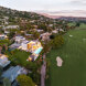 Vista aérea del atardecer de una villa contemporánea en primera línea de golf en Marbella, iluminada contra los exuberantes fairways verdes y ubicada al pie de las colinas, con vistas panorámicas a la montaña y un entorno residencial tranquilo.