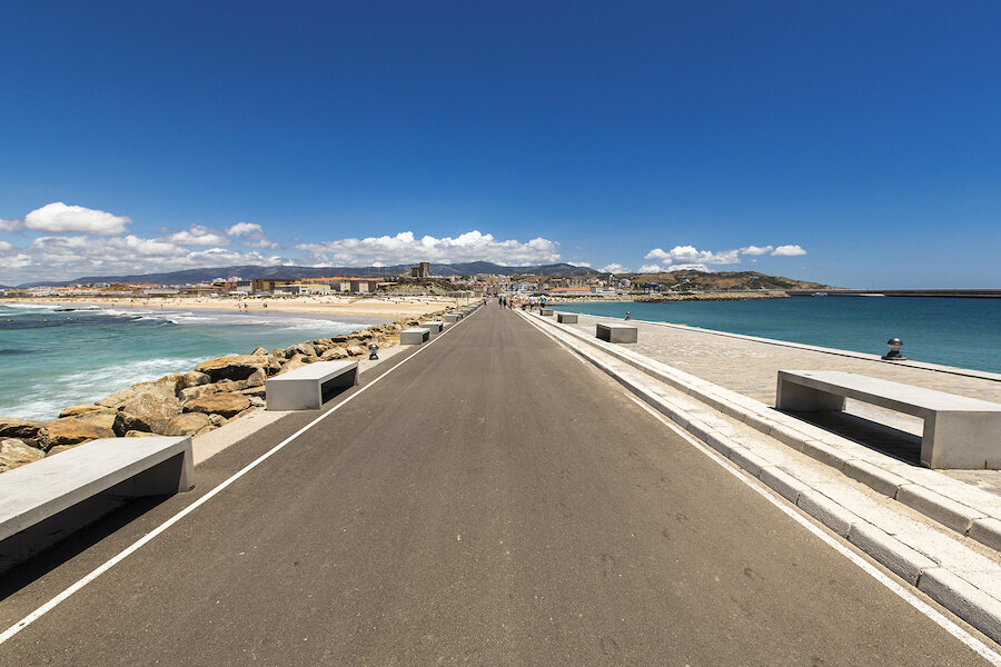 scenic escape. Looking towards Tarifa, a long paved walkway stretches into the distance, flanked by concrete benches. On the right side of the road is the Mediterranean Sea and on the left side is the Atlantic Ocean. In the background, the coastline, city buildings, and hills rise under a clear blue sky.