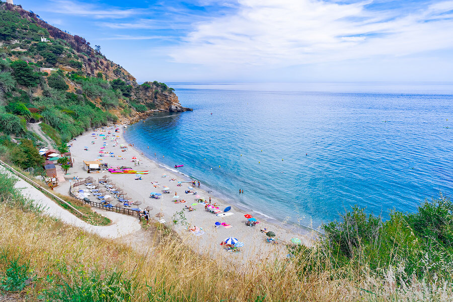 Eine friedliche Strandbucht an der Costa del Sol in Málaga mit Sonnenanbetern, bunten Sonnenschirmen, klarem blauen Wasser und üppig grünen Klippen, die die Küste unter einem leicht bewölkten Himmel umgeben.