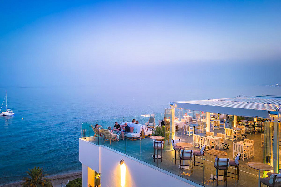 Marbella's fabulous rooftop bars. Elegant rooftop bar and restaurant overlooking the sea at dusk. Guests relax on sofas and dine at tables under a pergola, with soft lighting creating a cosy atmosphere. The calm water and a sailboat add to the serene coastal view.