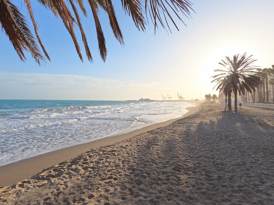 Sonnenuntergang an einem palmengesäumten Strand in Málaga, mit sanften Wellen, goldenem Licht und Silhouetten von Kränen in der Ferne in der Nähe des Hafens.
