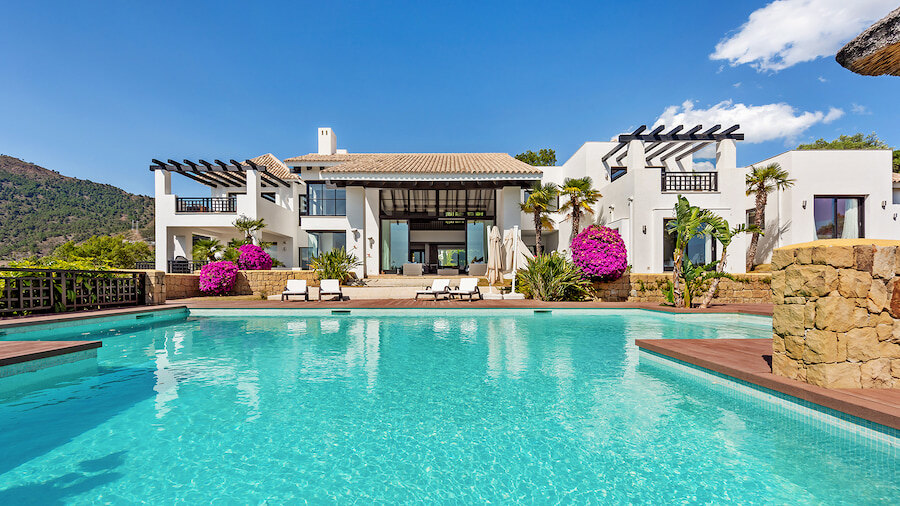 Vista frontal de una lujosa villa de estilo andaluz en La Zagaleta, Benahavís, que cuenta con una gran piscina infinita turquesa, buganvillas vibrantes y llamativos detalles arquitectónicos con un telón de fondo de cielos azules y exuberante vegetación.