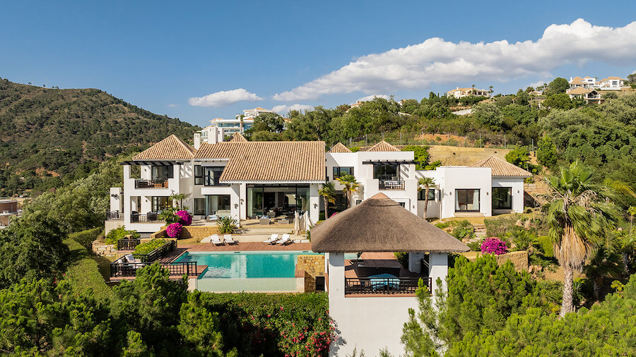 Vista aérea de una gran villa de lujo en La Zagaleta, Benahavís, con arquitectura tradicional andaluza, una gran piscina infinita, un cenador con techo de paja y hermosos jardines rodeados de exuberantes colinas y residencias exclusivas.