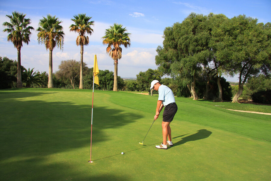 Golfista jugando en un green iluminado por el sol en un campo de golf de Marbella, rodeado de palmeras y exuberante vegetación, con cielos despejados y vistas panorámicas al fondo.