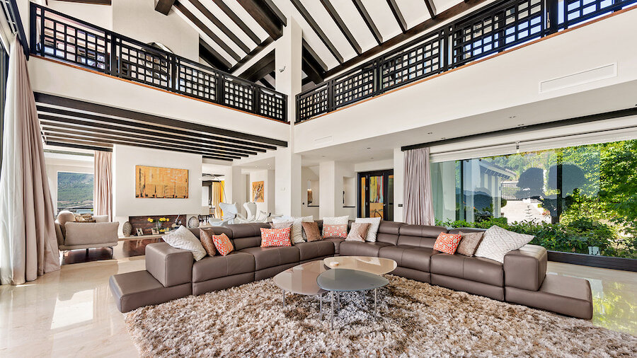 Amplia sala de estar de doble altura en una villa de lujo en La Zagaleta, con vigas de madera a la vista, una elegante galería en el entrepiso, grandes ventanales con vistas al jardín y una zona de estar central con una lujosa alfombra y muebles contemporáneos.