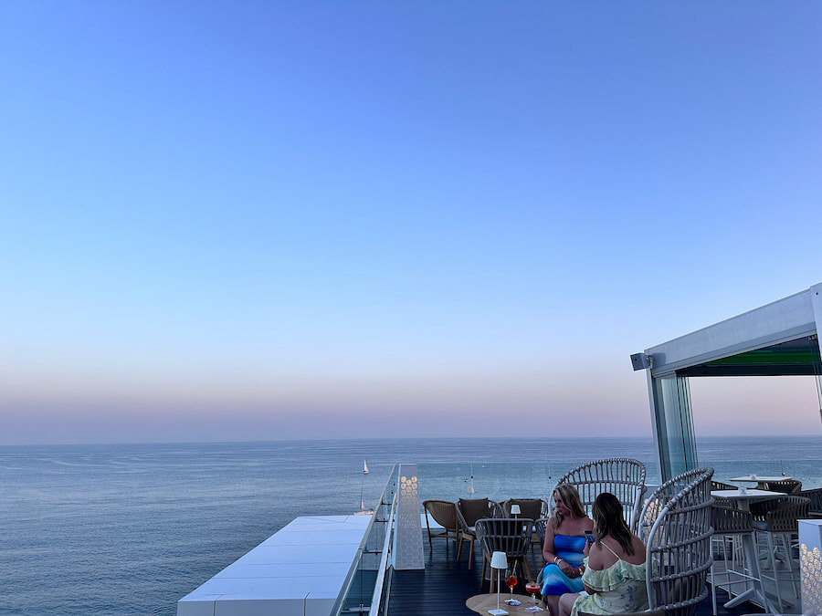 Two women enjoy cocktails on a rooftop terrace by the sea at sunset. The clear sky and calm water create a relaxing atmosphere, with wicker chairs and low tables in the foreground. In the distance, the horizon and a small sailboat can be seen.