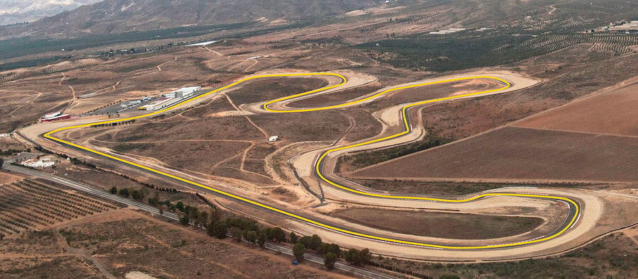 Luftaufnahme des Circuito de Almería, einer Rennstrecke, die sich durch eine trockene, wüstenähnliche Landschaft mit gelben Streckenmarkierungen schlängelt und von Hügeln, Ackerland und spärlicher Vegetation umgeben ist.
