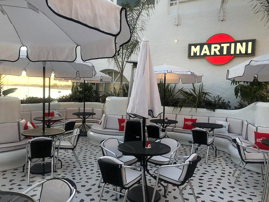 Chic outdoor seating area at Martini Bar by Dolce & Gabbana, featuring stylish black-and-white furniture, retro umbrellas, and red branded cushions. The iconic Martini logo glows on the wall, adding a glamorous touch to the elegant, Mediterranean-inspired setting.