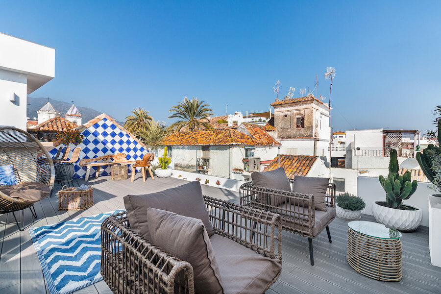 Sunny rooftop terrace at Linda Boutique Hotel Marbella, featuring wicker lounge chairs, a blue zigzag rug, and a dining area with a traditional tiled wall. Surrounded by charming old town rooftops, potted plants, and mountain views in the background.
