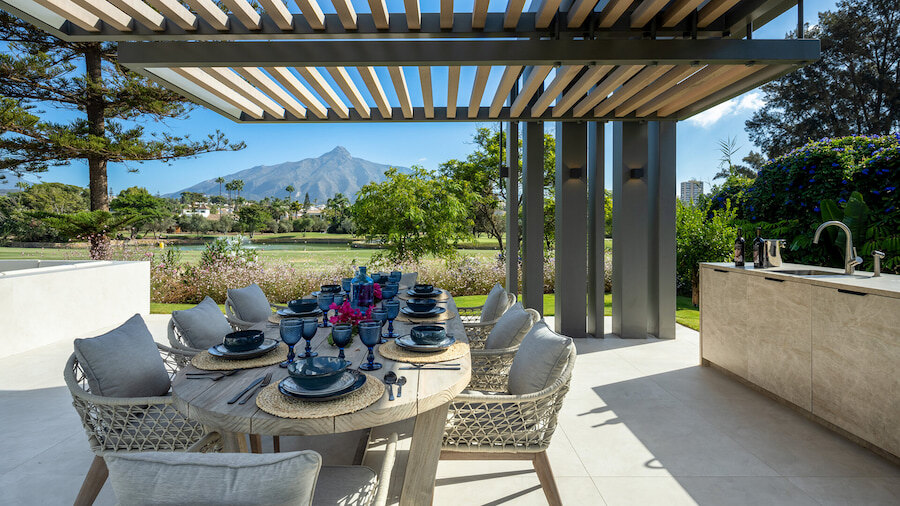 Elegante comedor al aire libre en una villa de lujo de Marbella, con pérgola cubierta, cocina de verano incorporada y una mesa bellamente dispuesta con vistas al campo de golf y a la montaña La Concha al fondo.