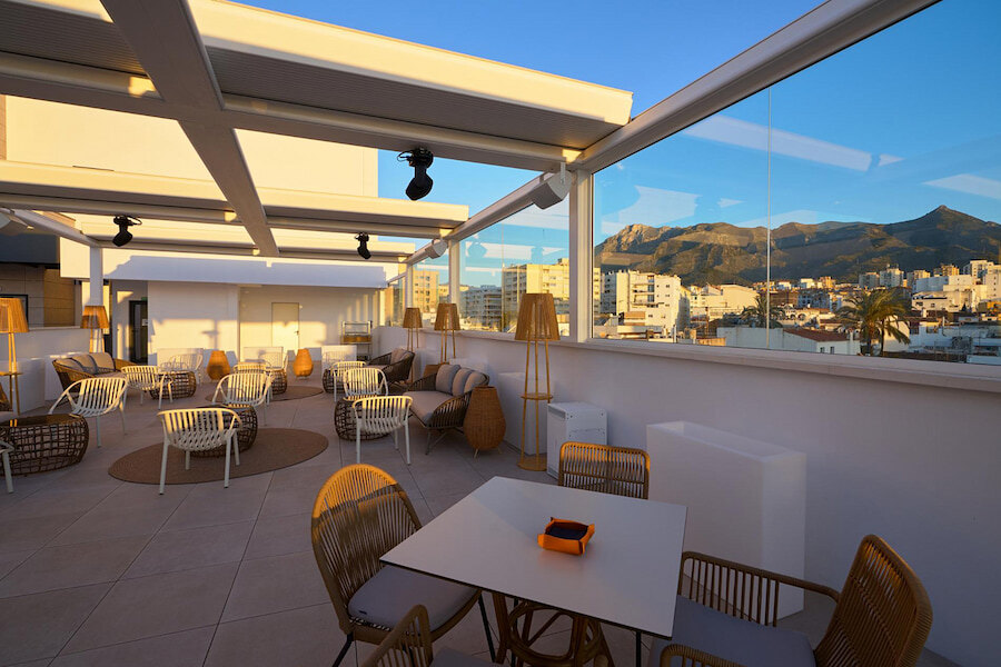 Stylish rooftop terrace with wicker chairs and modern decor, overlooking the city and surrounding mountains at golden hour. The glass walls offer panoramic views, while the warm sunlight creates a cosy and inviting atmosphere.