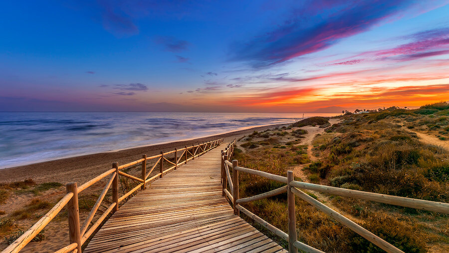 Holzsteg, der bei Sonnenuntergang zu einem wilden Strand in Málaga führt, mit farbenfrohem Himmel, Küstendünen und Wellen, die vom Meer heranrollen.