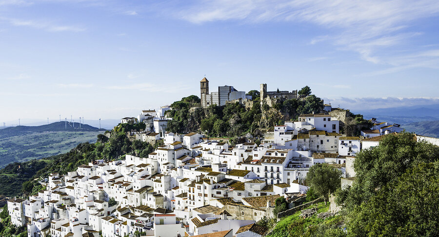 Panoramic view of a traditional Andalusian white village perched on a hillside, with a church tower and castle ruins at the top. Rolling green hills and distant wind turbines stretch across the horizon under a partly cloudy sky.