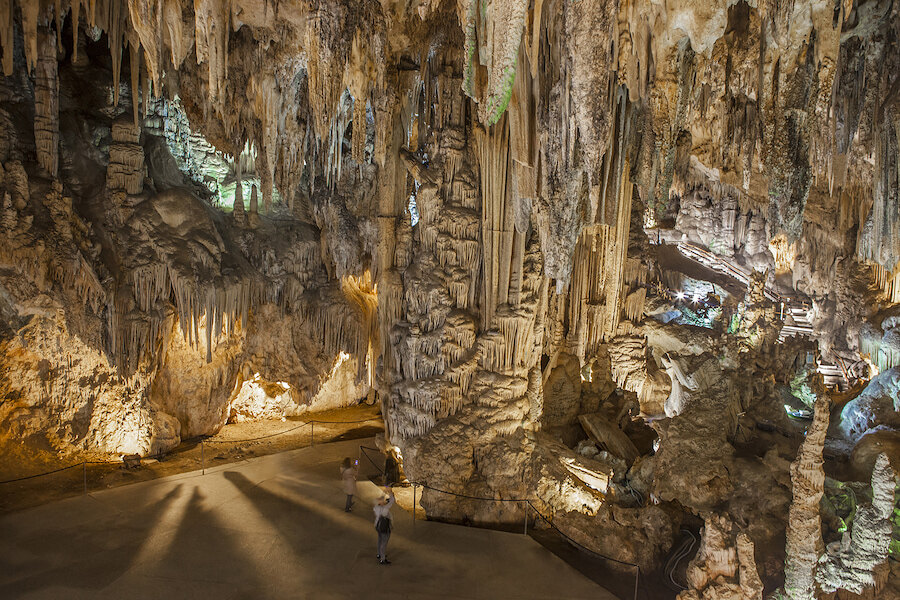 Interior view of an illuminated cave with impressive stalactites and stalagmites. Visitors walk along a designated path, dwarfed by the massive rock formations in this natural underground wonder.